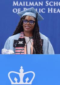 A young Black woman with long hair and glasses speaks at a podium wearing blue cap and gown
